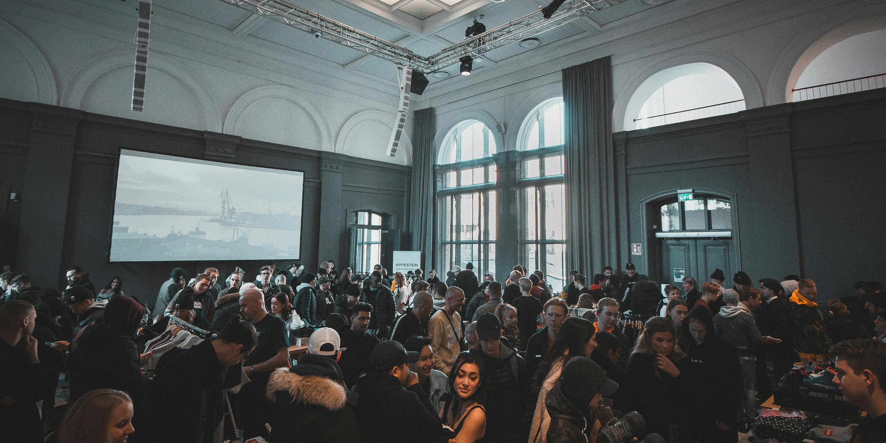 A bustling event venue with a large crowd, tall windows, and a projection screen showing an industrial scene.