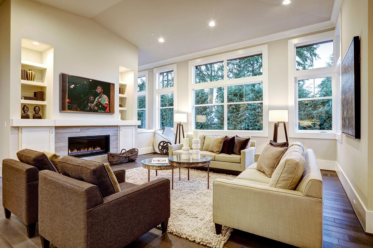 Bright, spacious living room with multiple sofas and a wall-mounted TV above a fireplace, surrounded by large windows.