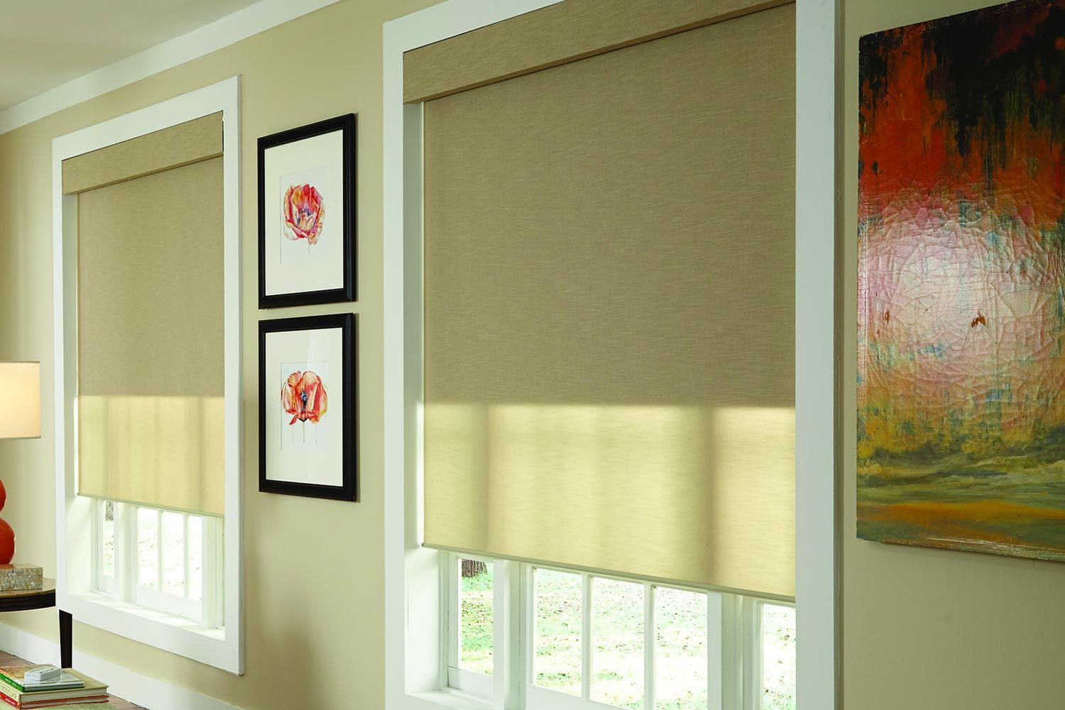 A warm-toned room with two large windows featuring beige blackout shades, framed art pieces, and a vibrant painting on the wall.