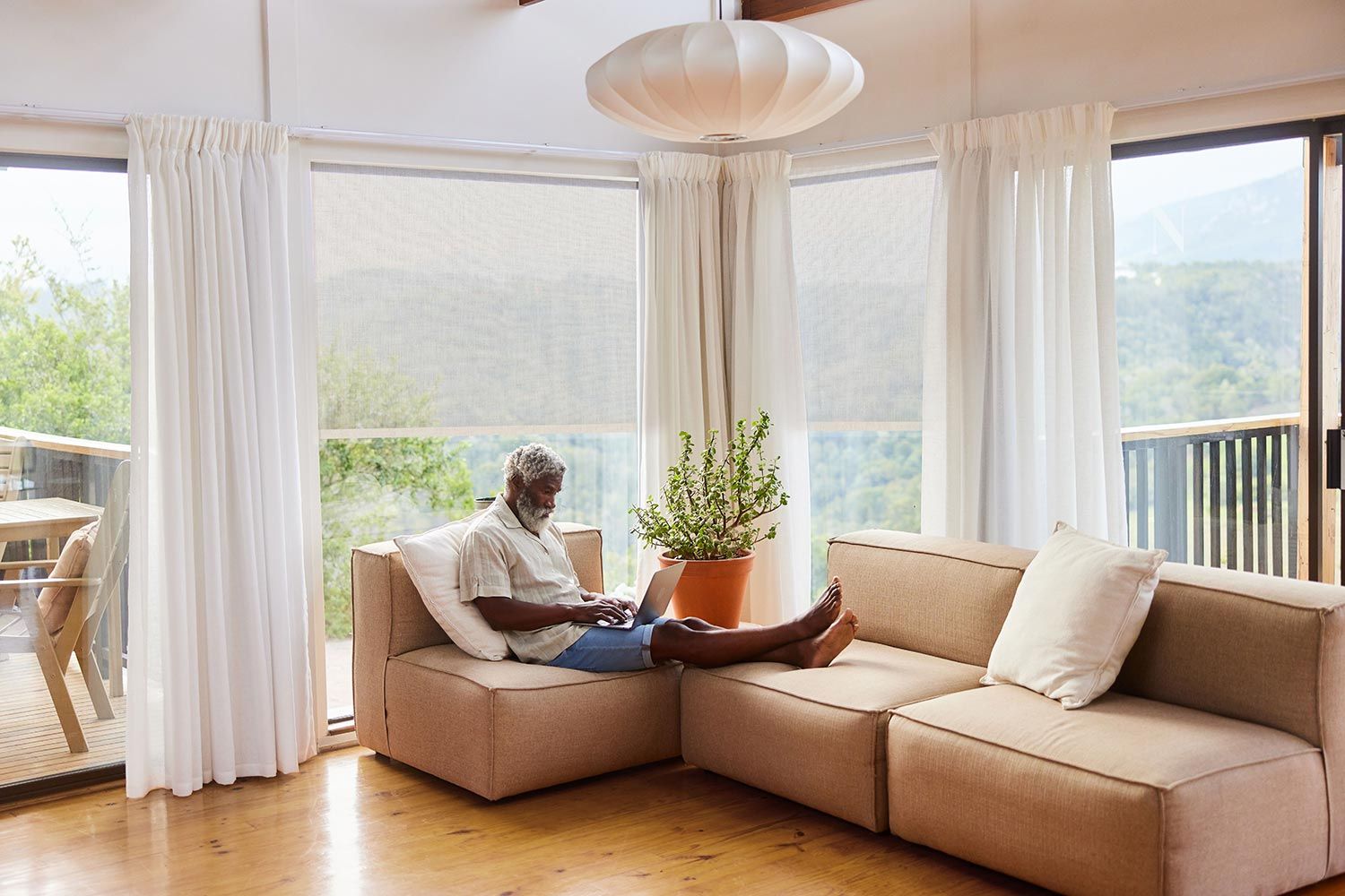 A serene living room with a man relaxing on a sofa with a laptop, surrounded by natural light and large windows with curtains.