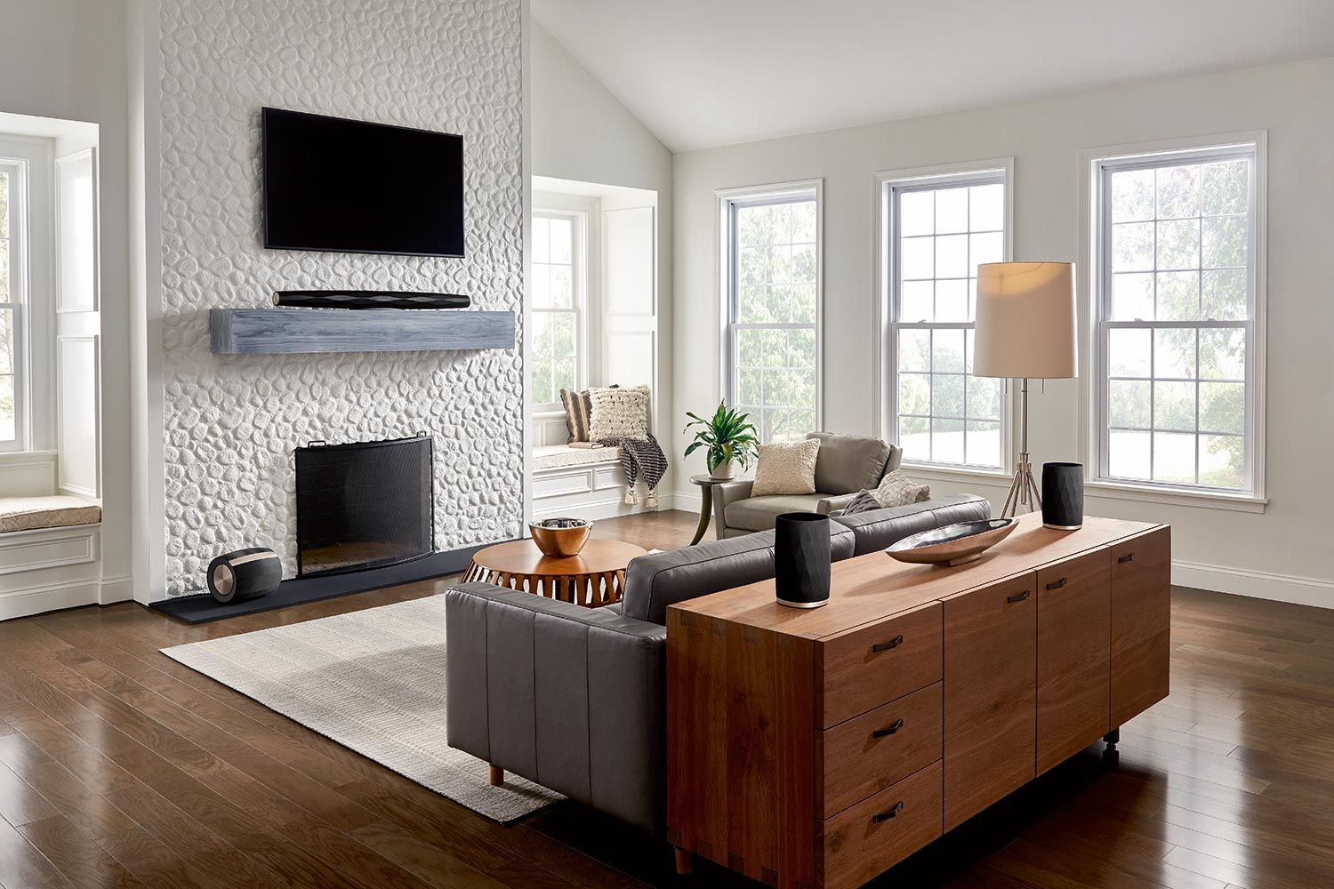 A bright and airy living room with a mounted TV above a textured stone fireplace, a modern sofa, and several windows bringing in natural light, featuring audio equipment placed discreetly around the room.