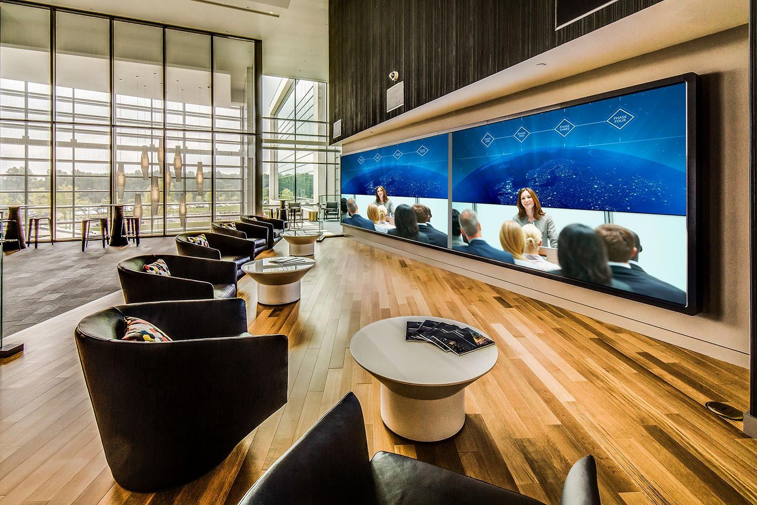 A sophisticated lounge area with leather chairs, large video walls displaying presentations, and floor-to-ceiling windows offering a scenic view.