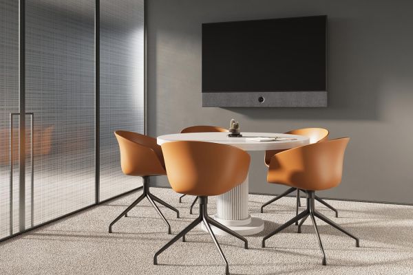 A small, modern meeting room with a round table, orange chairs, and a wall-mounted display, designed for intimate discussions.