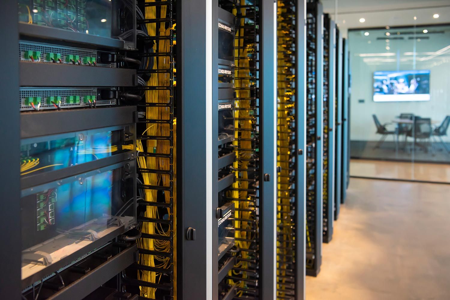 Server room with organized racks of networking equipment and neatly bundled yellow cables, visible meeting room in the background.